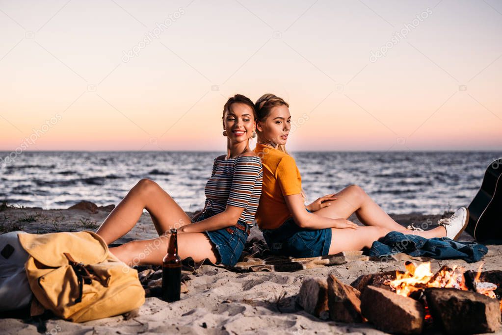 happy young women sitting back to back on sandy beach at sunset