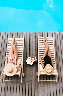 high angle view of young women in swimwear and hats lying on chaise lounges near swimming pool  clipart