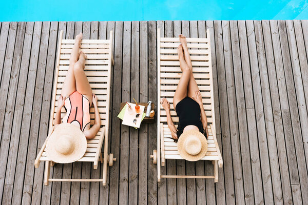 молодые женщины в купальниках и шляпах, лежащие в шезлонгах возле бассейна
 