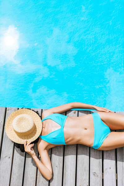 Вид сверху на молодую женщину в бикини и соломенной шляпе на лице, лежащую возле бассейна
 