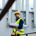 Engenheiro masculino em colete de segurança e capacete com planta usando tablet digital na construção