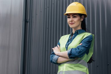 Emanet yelek ve kask ayakta duvara çapraz kolları olan kendine güvenen kadın mimar