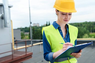 beautiful female engineer in safety vest and hardhat writing in clipboard clipart