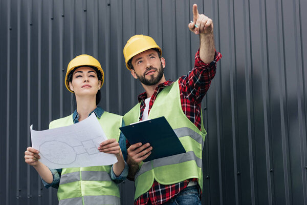 architects in helmets pointing and working with blueprint and clipboard on roof
