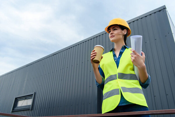 beautiful female engineer in safety vest and helmet with blueprint and disposable cup on coffee