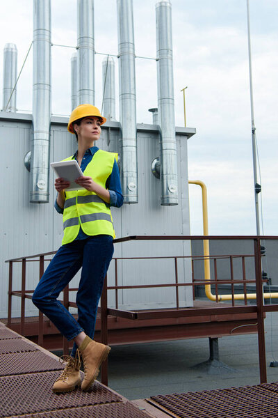 female architect in safety vest and hardhat using digital tablet on industrial construction