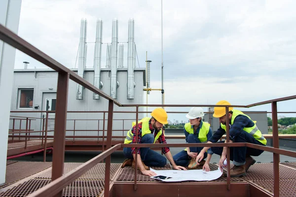 三名工程师在头盔上与屋顶上的蓝图一起工作 — 图库照片