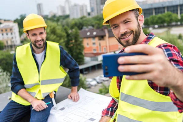 Ingenieros Masculinos Chalecos Seguridad Cascos Con Planos Tomando Selfie Teléfono —  Fotos de Stock