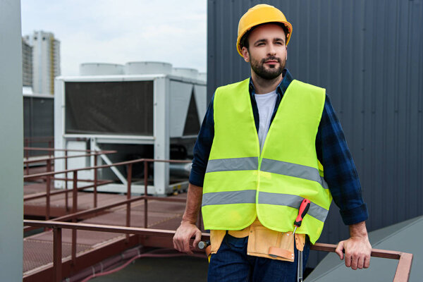 male architect in safety vest and hardhat with tool belt standing on roof 