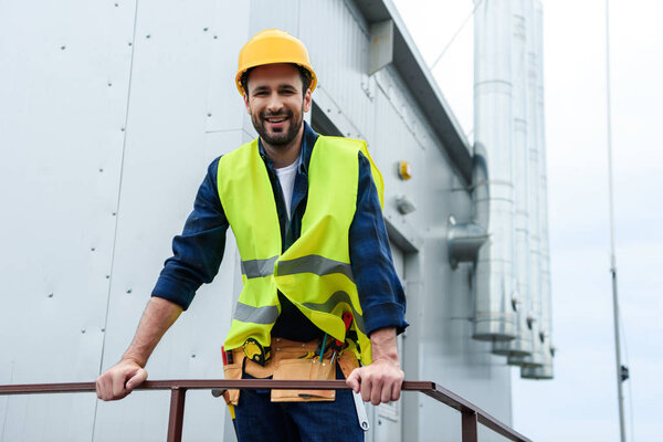 male smiling engineer in safety vest and helmet with tool belt on construction