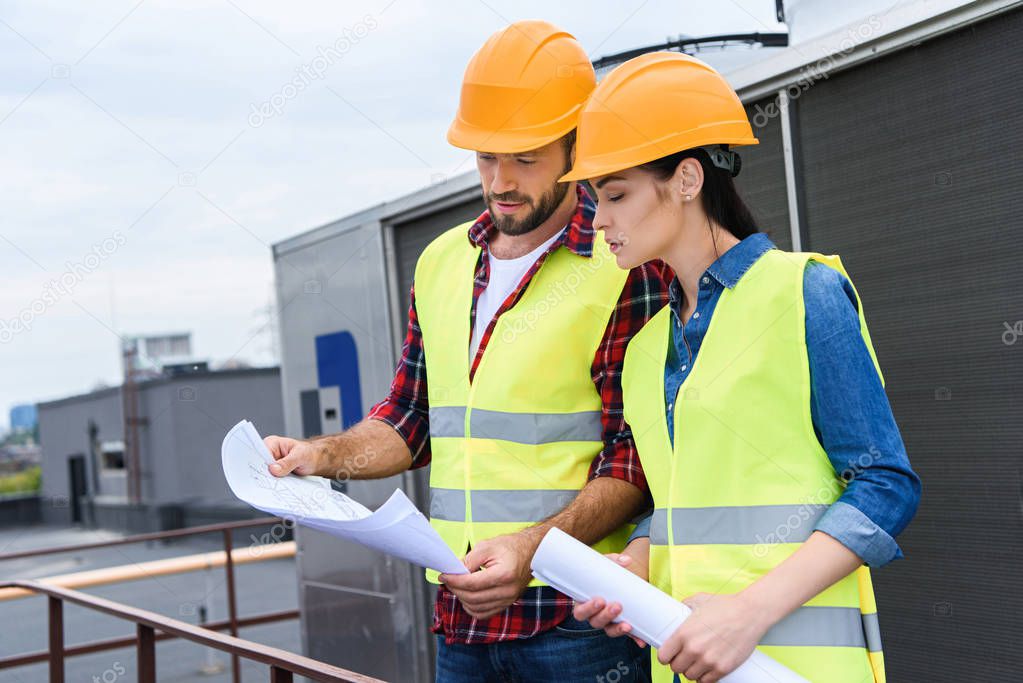 professional architects in helmets working with blueprints on roof