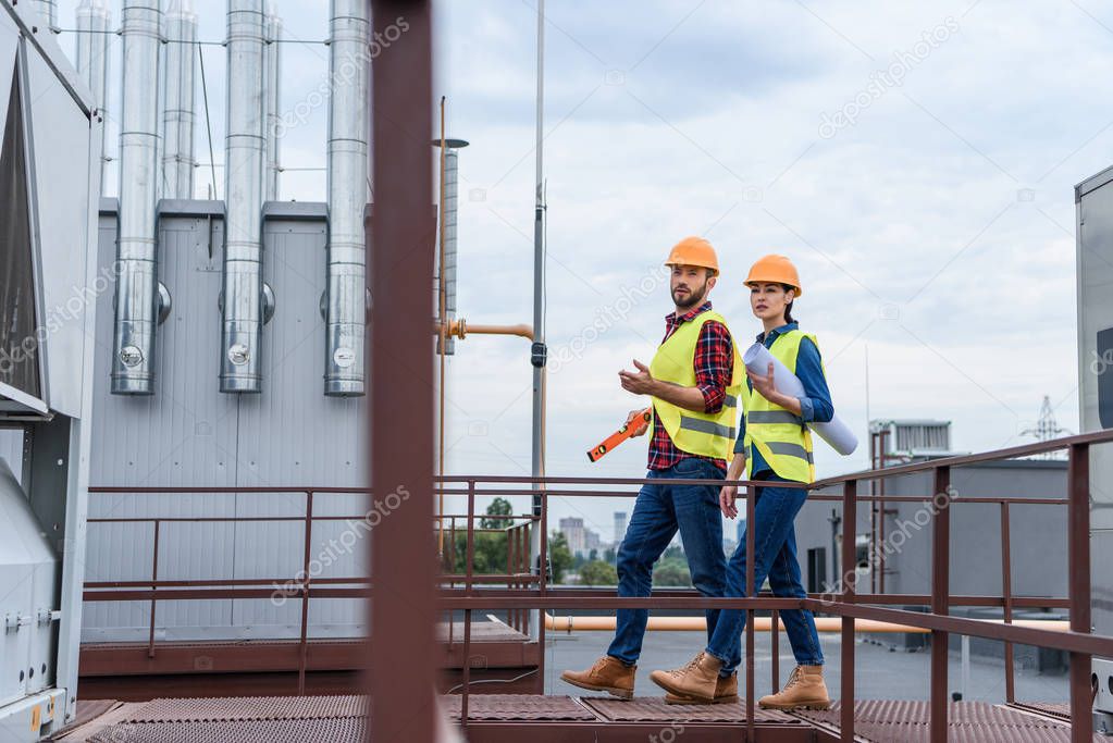 professional architects in helmets with blueprint and level walking on roof