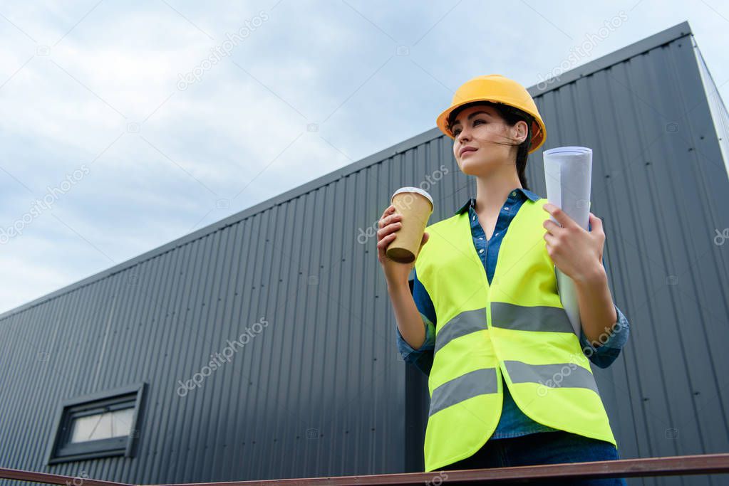 beautiful female engineer in safety vest and helmet with blueprint and disposable cup on coffee