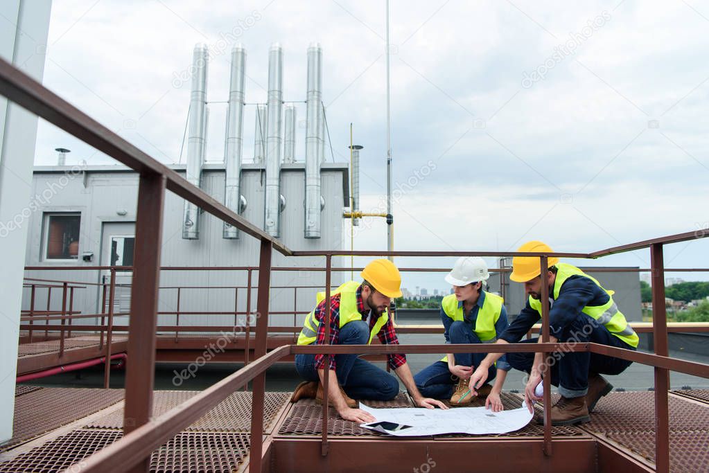 three engineers in helmets working with blueprints on roof together