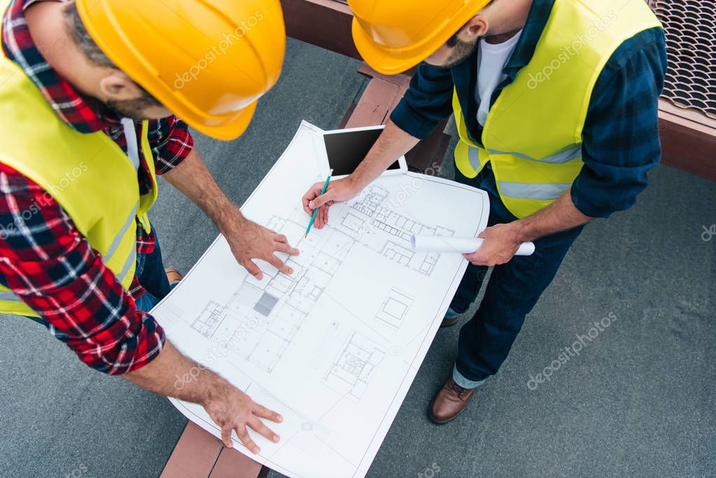 overhead view of architects in safety vests and helmets drawing on blueprints