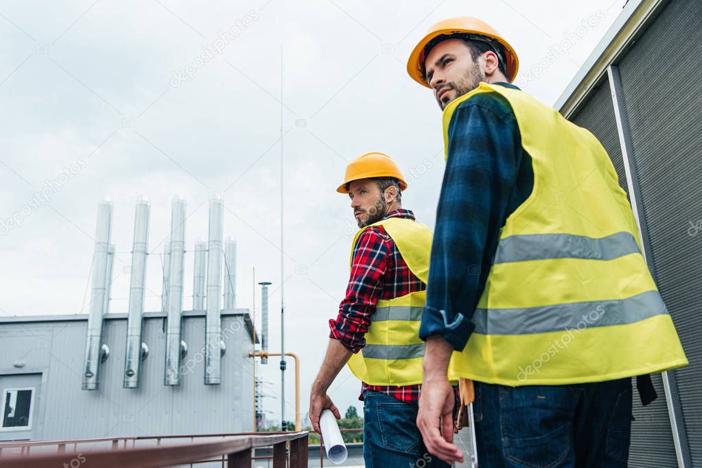 smiling engineers in safety vests and helmets with blueprint on roof