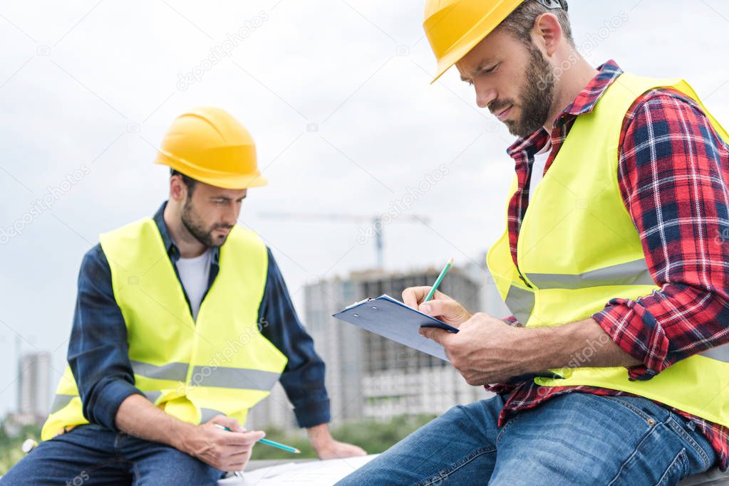 two engineers in safety vests and hardhats working with blueprints and clipboard on roof
