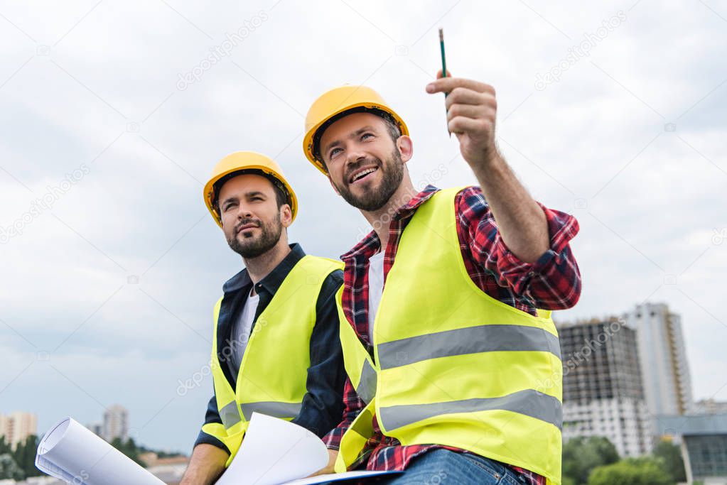 two architects in safety vests and hardhats working with blueprints and pointing at construction