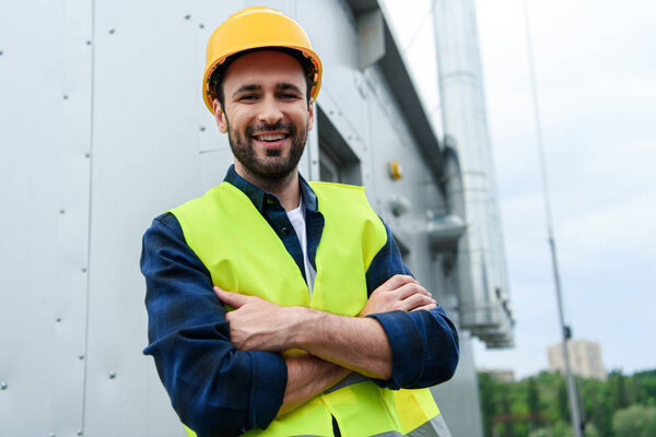 male smiling engineer in safety vest and helmet standing with crossed arms