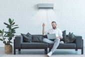 schöner Mann schaltet Klimaanlage mit Fernbedienung ein, während er Laptop auf dem heimischen Sofa benutzt