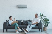 Paar schaltet Klimaanlage in der Sommerhitze ein, während es mit Buch und Laptop auf dem Sofa sitzt 