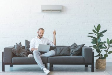 man turning on air conditioner with remote control while using laptop on sofa clipart