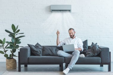 handsome man turning on air conditioner with remote control while using laptop on sofa at home clipart