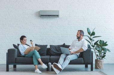 couple turning on air conditioner during the summer heat while sitting on sofa with book and laptop  clipart