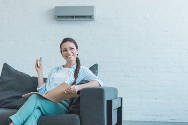smiling woman turning on air conditioner with remote control while reading book on sofa clipart