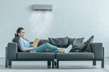 woman reading book on couch, air conditioner blowing on her