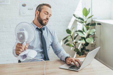 işadamı kendini ofiste dizüstü bilgisayar kullanırken Elektrikli fan ile soğutma