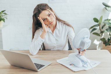 upset businesswoman sitting at workplace with paperwork, laptop and electric fan 
