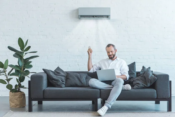 Homem Bonito Ligando Condicionado Com Controle Remoto Usar Laptop Sofá — Fotografia de Stock