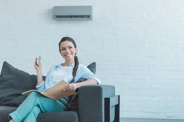 Mujer Sonriente Encendiendo Aire Acondicionado Con Control Remoto Mientras Lee —  Fotos de Stock