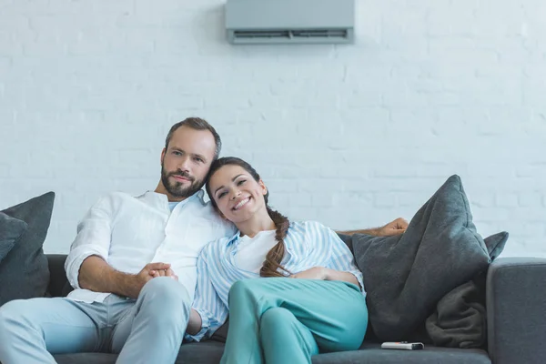 smiling couple sitting on sofa during the summer heat at home with air conditioner on wall