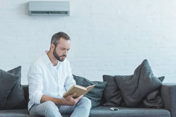 Bebaarde Man Lezen Boek Bank Met Air Conditioner Muur — Stockfoto