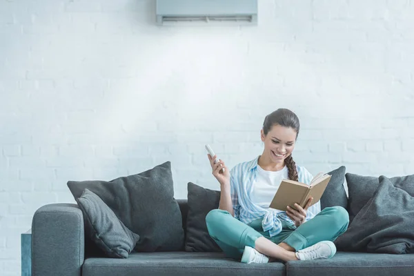 Happy Woman Reading Book While Turning Air Conditioner Remote Control — Stock Photo, Image