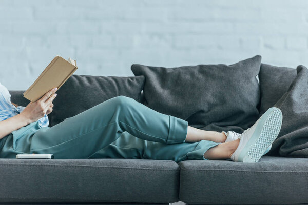 cropped view of woman relaxing on sofa and reading book 