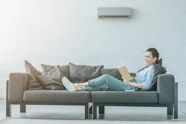 Happy Woman Reading Book Couch Air Conditioner Wall Summer Heat — Stock Photo, Image