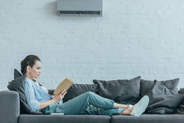 woman reading book on sofa, with air conditioner on wall at home