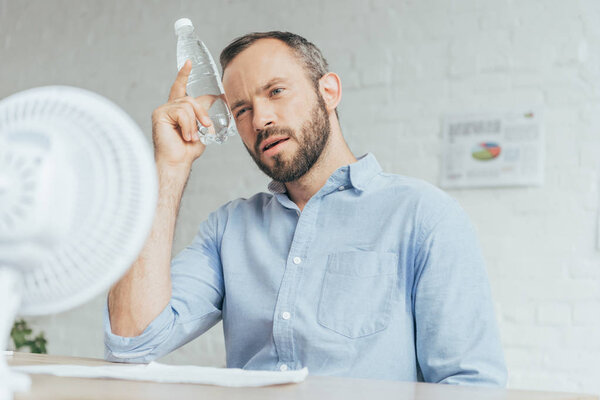 businessman cooling himself with bottle of water and conditioning air with electric fan in office