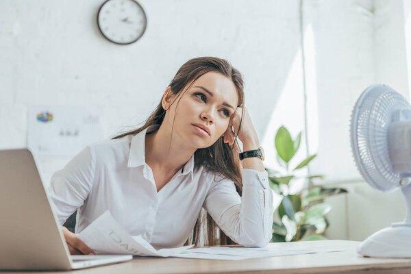 tired businesswoman sitting at table with paperwork, laptop and electric fan