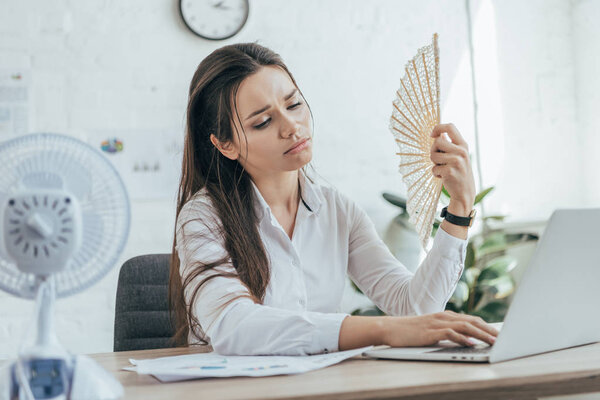 exhausted businesswoman using laptop while conditioning air with electric fan and hand fan in office