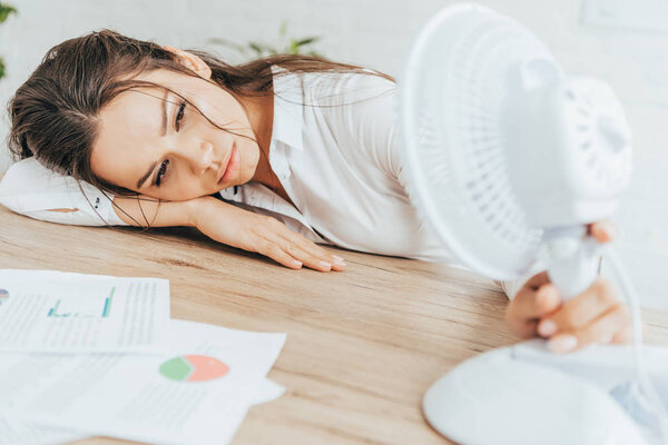 exhausted businesswoman blowing at herself with electric fan while lying on table with documents