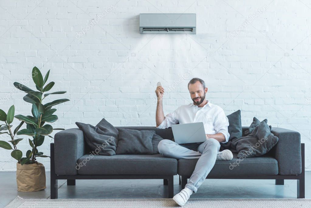 Handsome man turning on air conditioner with remote control while using laptop on sofa at home