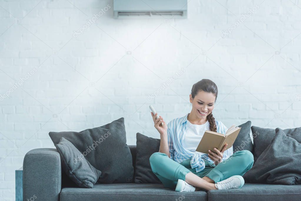 happy woman reading book while turning on air conditioner with remote control at home