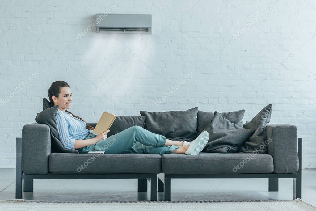 Woman reading book on couch, air conditioner blowing on her