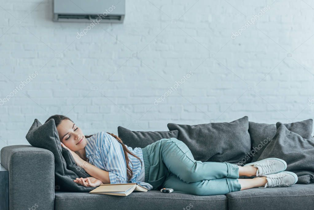 Woman sleeping on sofa with book and air conditioner on wall
