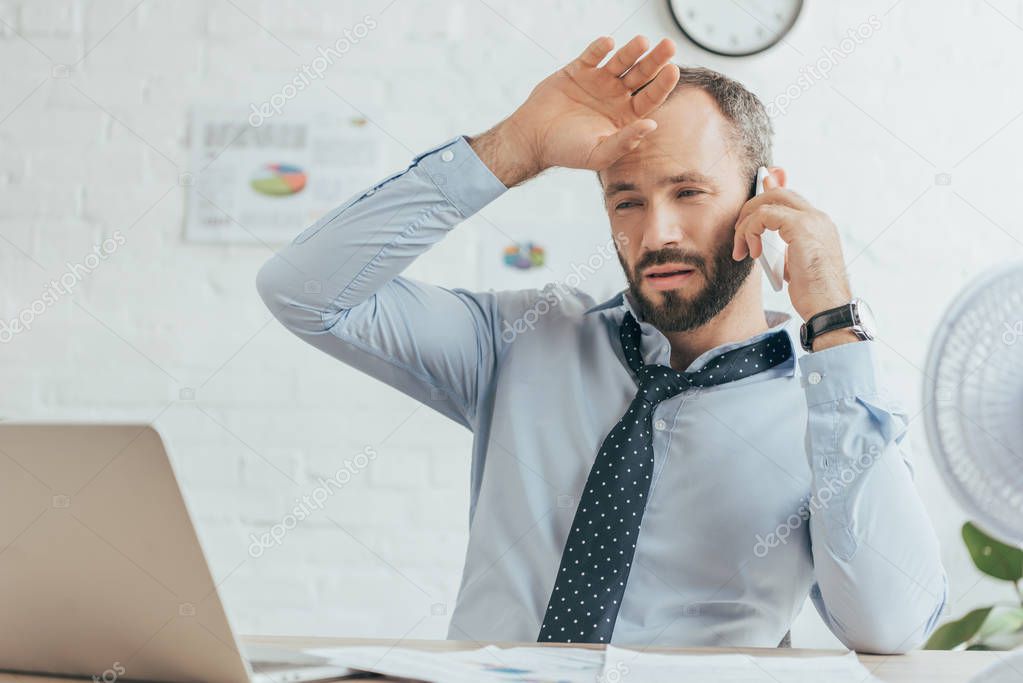 exhausted sweaty businessman talking on smartphone and working in hot office