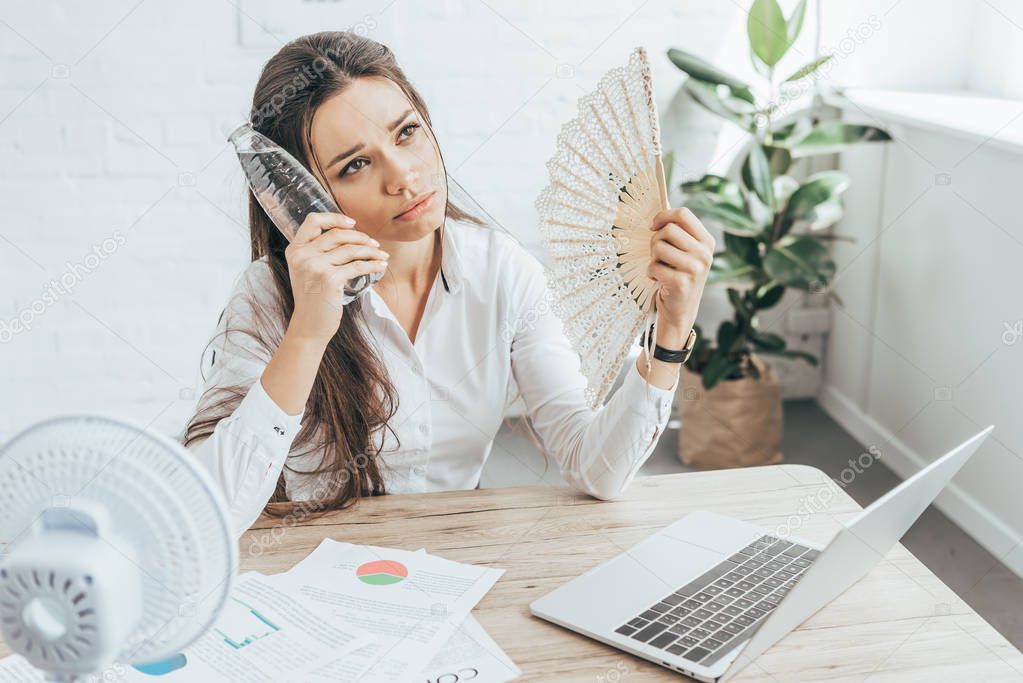 businesswoman cooling herself with electric fan, hand fan and bottle of water at workplace with documents and laptop